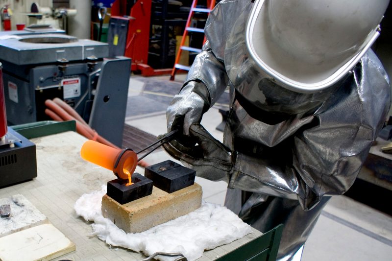 Inside the Kroehling foundry, a person wearing protective equipment and a helmet has tongs holding a red hot crucible containing molten gold which is pouring into a ingot mold.
