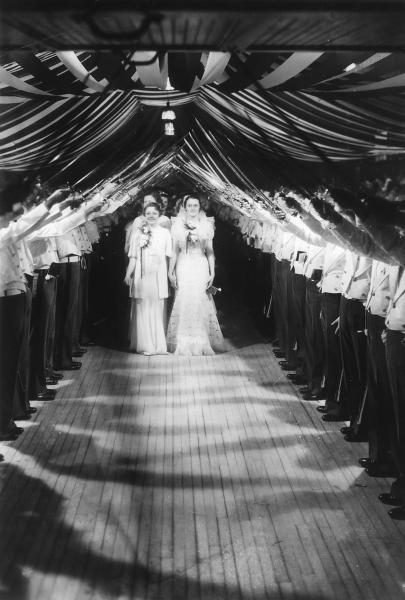 Black and white photo. A sabre arch by cadets with a line of women in white dresses ready to process under the arch.