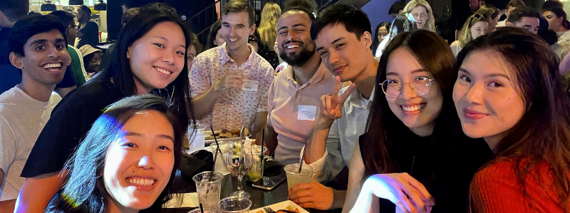 group of young alumni smiling and sitting around a table with drinks