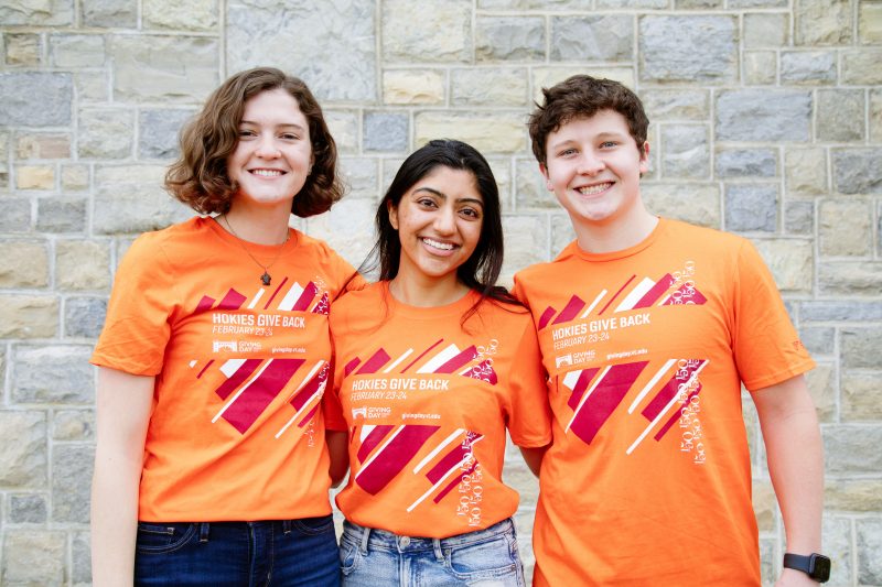 Three students, female, female, male standing together in front of Hoke stone wall, wearing orange Giving Day t-shirts.