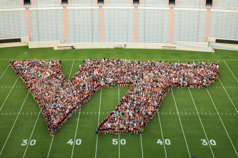 Members of Class of 2025 form a large outline of the athletic VT centered on green Worsham Football Field at Lane Stadium. Empty East stands are in background, numbered yardlines 30, 40, 50, 40, 30 at bottom of photo.