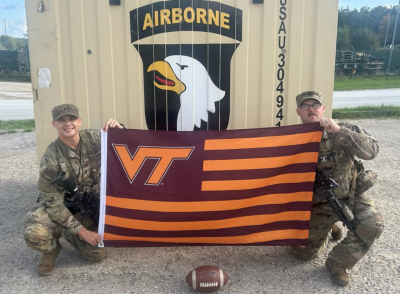 Two smiling servicemen in camouflage uniforms kneel while holding a Virginia Tech flag. The artwork on the structure behind them says “Airborne”. 