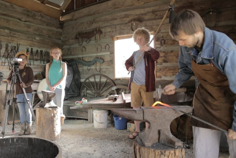 Three children watch as a blacksmith bends a glowing orange piece of metal. Once child films the blacksmith with a video camera, while another holds a microphone up over the blacksmith. A third child in the middle watches the blacksmith work. 