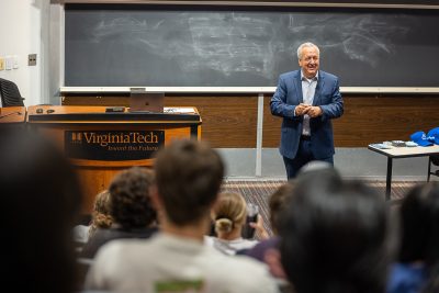 A man presenting in front of a room of individuals as viewed from the back of the room through blurred heads.